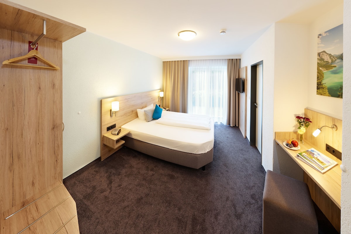 A hotel room with a bed, a desk, a TV, and a painting on the wall.