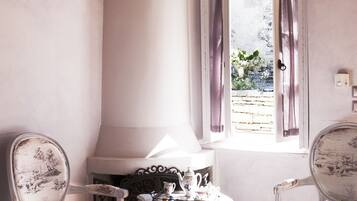 A room with a fireplace, two chairs, a small table with a teapot, and a window with curtains.