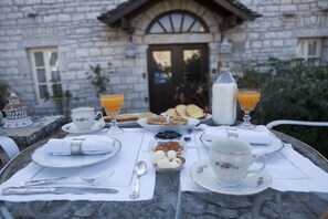 Une table dressée pour le petit-déjeuner, avec en arrière-plan un bâtiment en pierre, des assiettes, des tasses et un verre de jus d’orange.