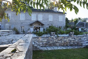 Un bâtiment en pierre doté d’une entrée centrale, d’un escalier en pierre menant à l’entrée et d’une terrasse pavée aménagée avec du mobilier d’extérieur.
