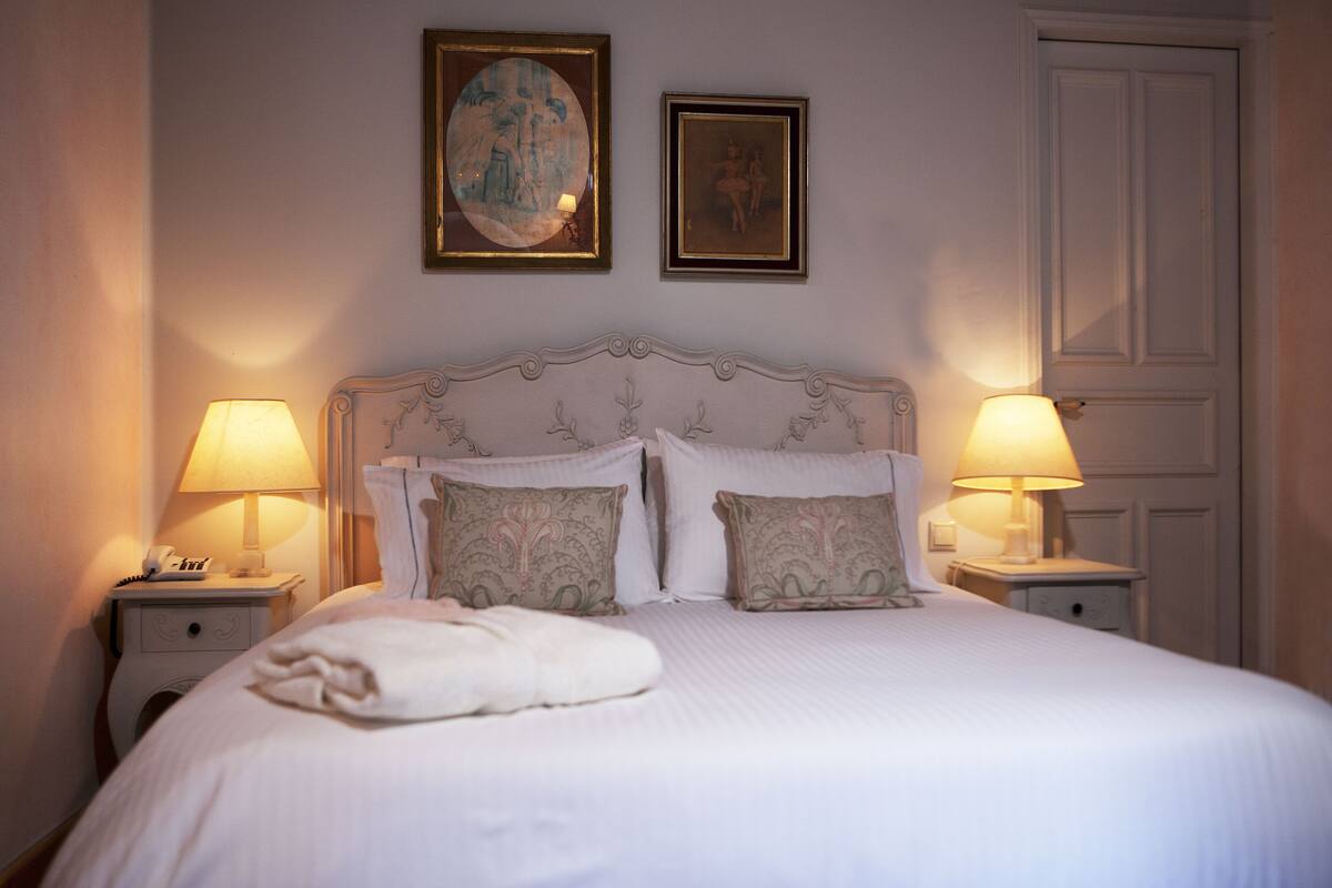 A neatly made bed with a white comforter and decorative pillows, two bedside lamps, and a framed artwork above the bed.