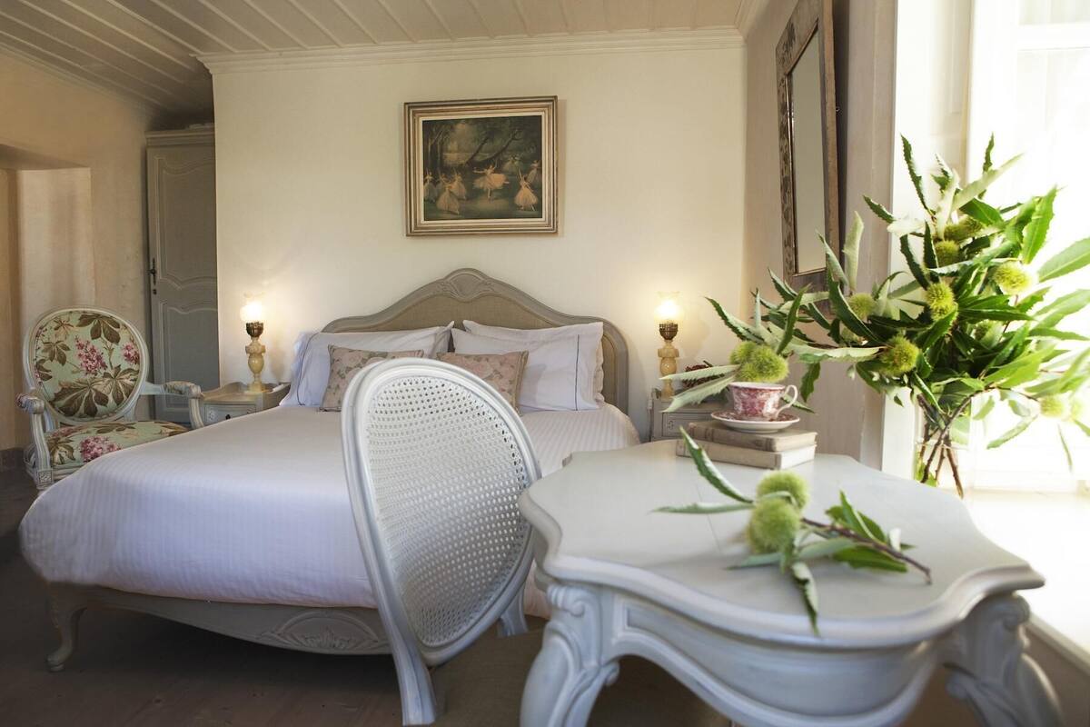 A bedroom with two beds, a chair, a table with a plant, and a painting on the wall.