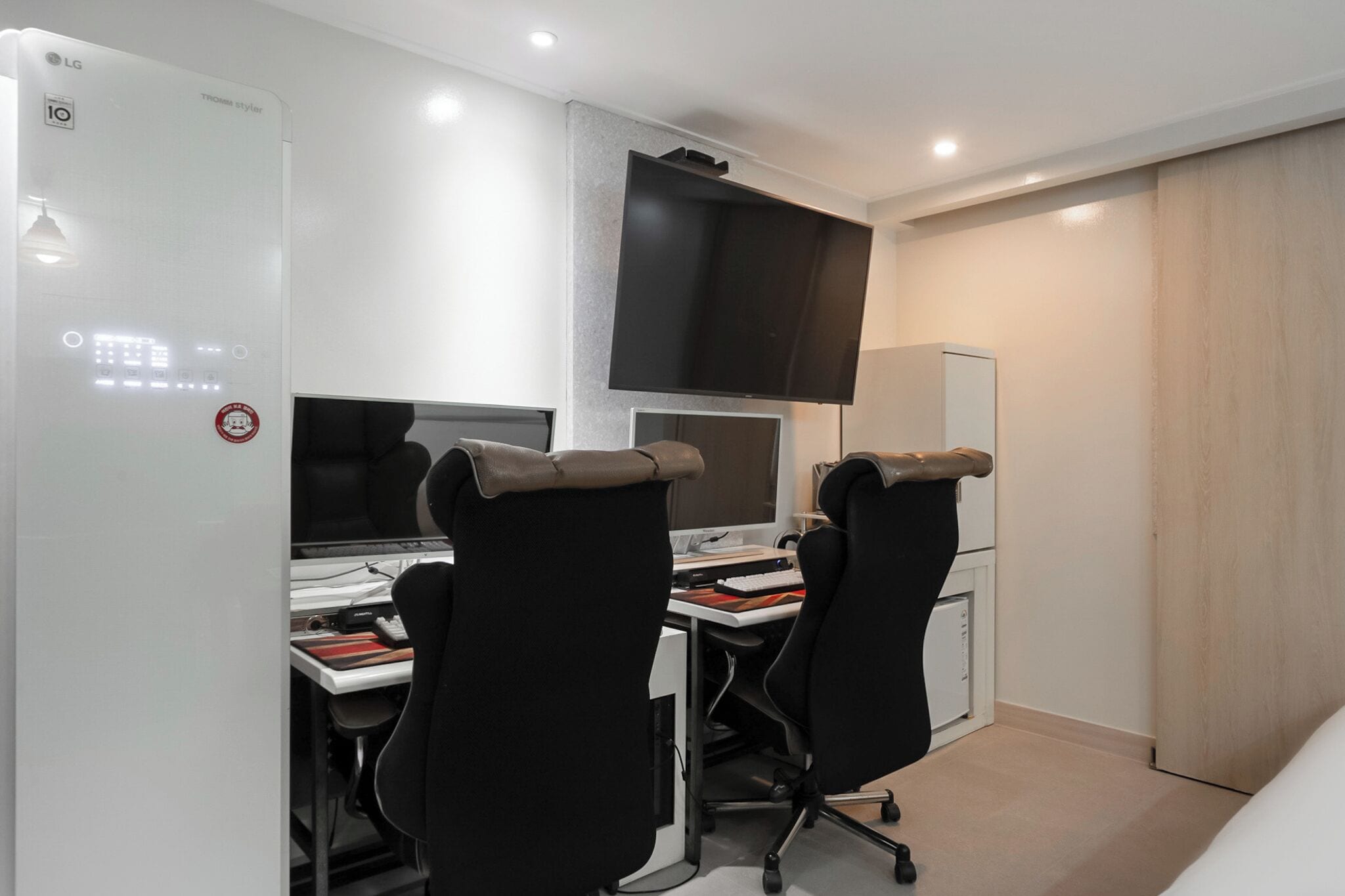 A modern office space with a white desk, two black office chairs, a flat-screen monitor, a keyboard, a mouse, and a large flat-screen TV mounted on the wall.