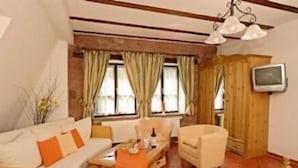 A cozy room with a sofa, armchair, and a small table with a bottle and glasses. There is a television mounted on the wall and a window with curtains. - Zum Alten Wasserrad - L Antica Ruota (Annweiler am Trifels)