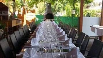 A long dining table set for a formal meal with chairs on both sides, under a wooden pergola with a view of greenery.