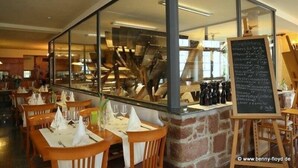 A restaurant with wooden tables and chairs, a glass partition, and a chalkboard menu. - Zum Alten Wasserrad - L Antica Ruota (Annweiler am Trifels)