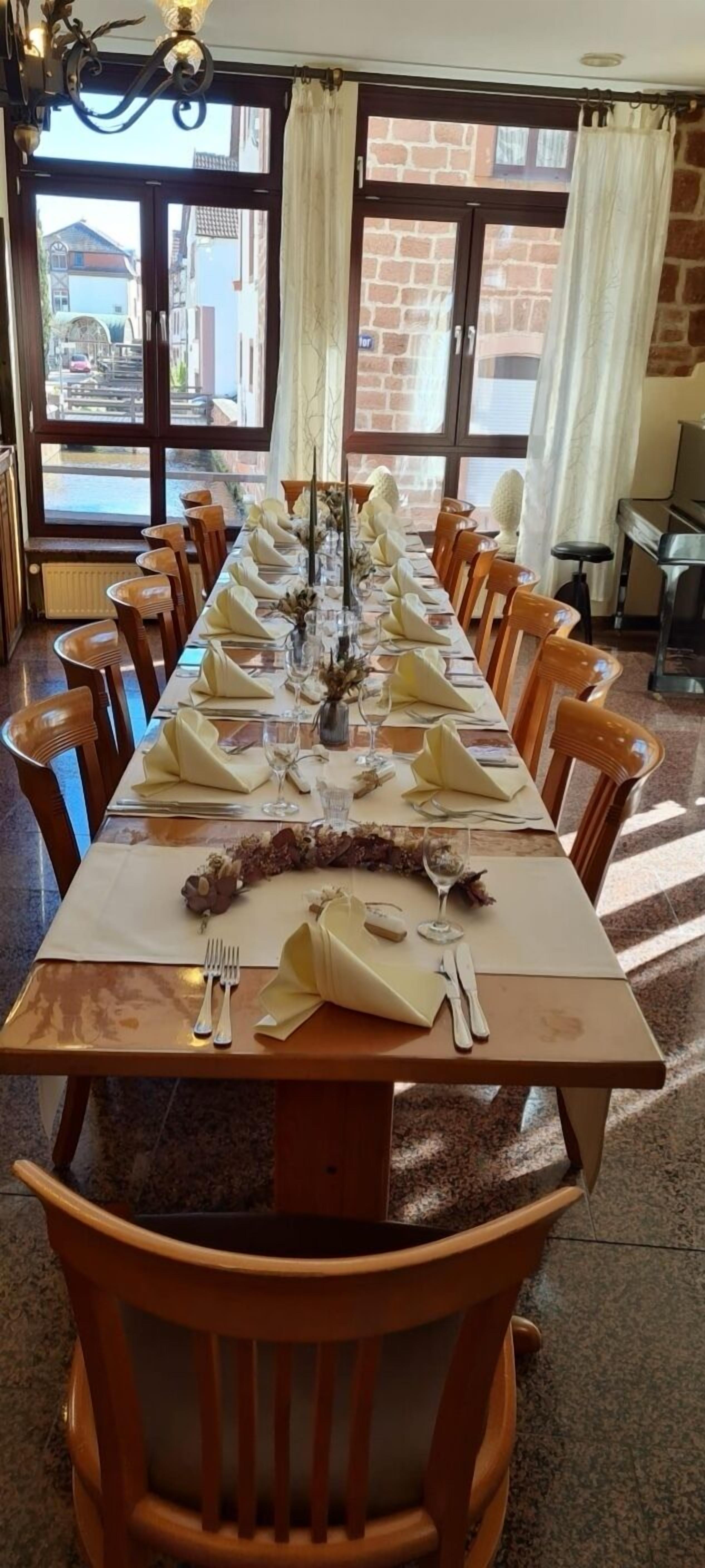Una lunga tavola da pranzo apparecchiata con sedie, un lampadario e vista sulla strada attraverso ampie finestre.