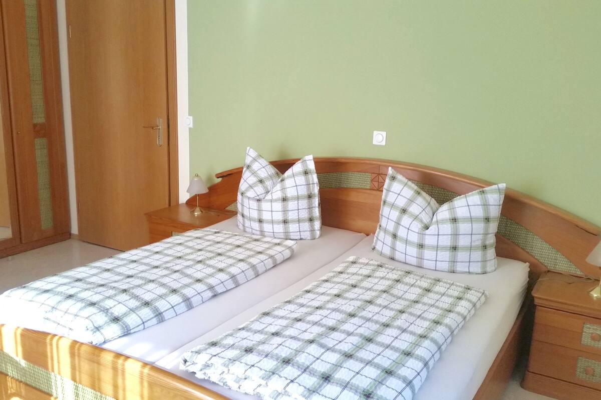 Uma cama de casal com colcha xadrez e almofadas a combinar, cabeceira de madeira, mesa de cabeceira com abajur e guarda-roupa de madeira com espelho.