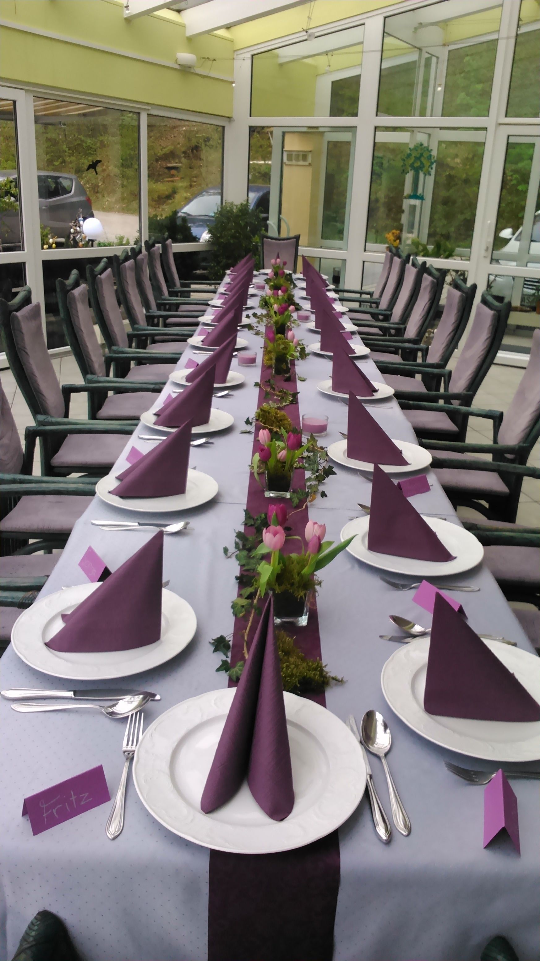 Una larga mesa de comedor dispuesta con servilletas moradas, platos blancos y cubiertos, rodeada de sillas en una habitación luminosa e inundada de luz solar con ventanas amplias.