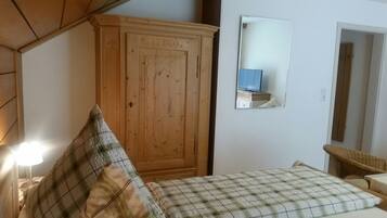 A bedroom with a wooden wardrobe, a bed with plaid bedding, a television on a stand, and a wicker chair.