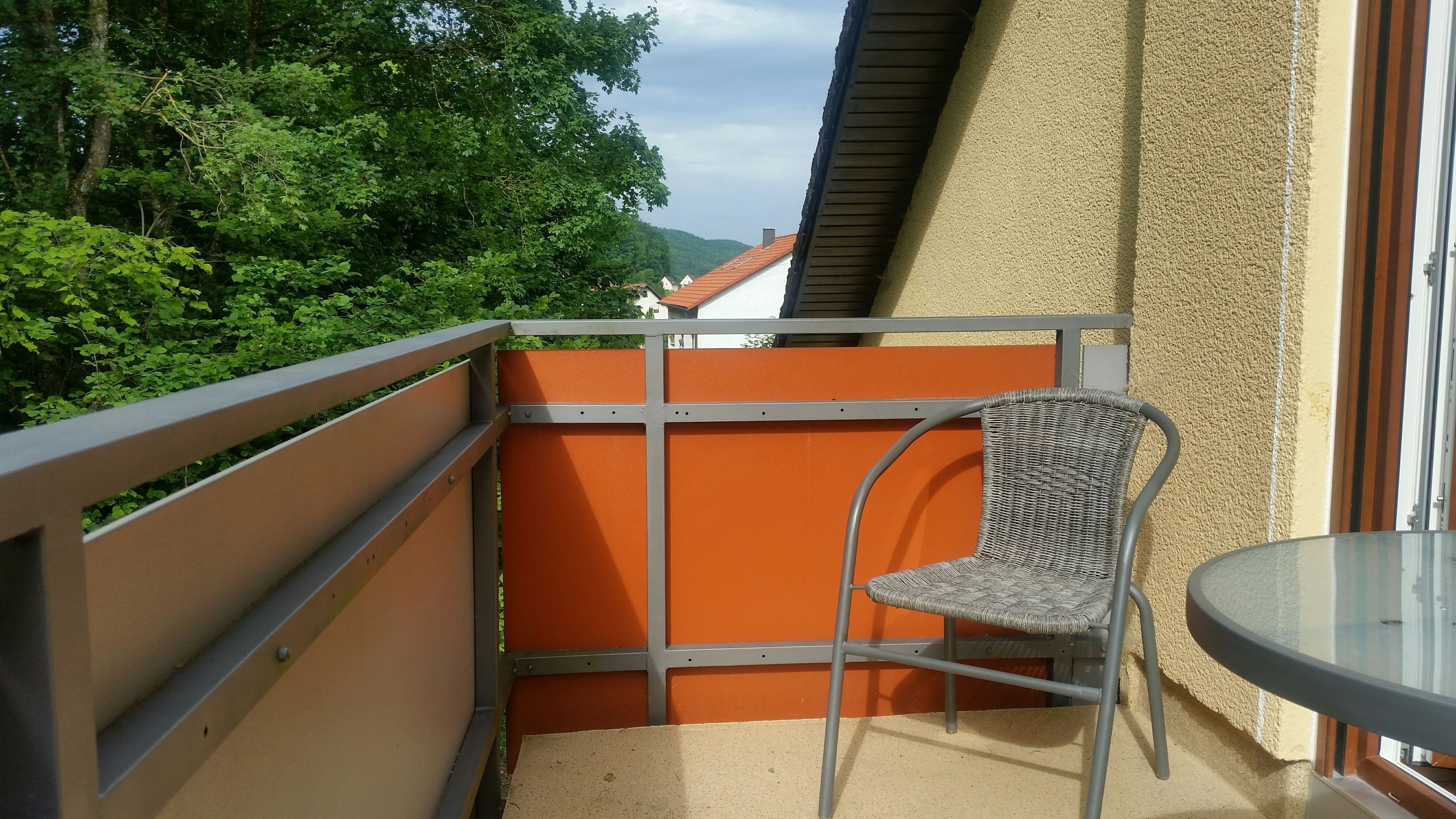 Ein Balkon mit Korbstuhl und kleinem Tisch, umgeben von einem Metallgeländer und orangefarbenen Paneelen.