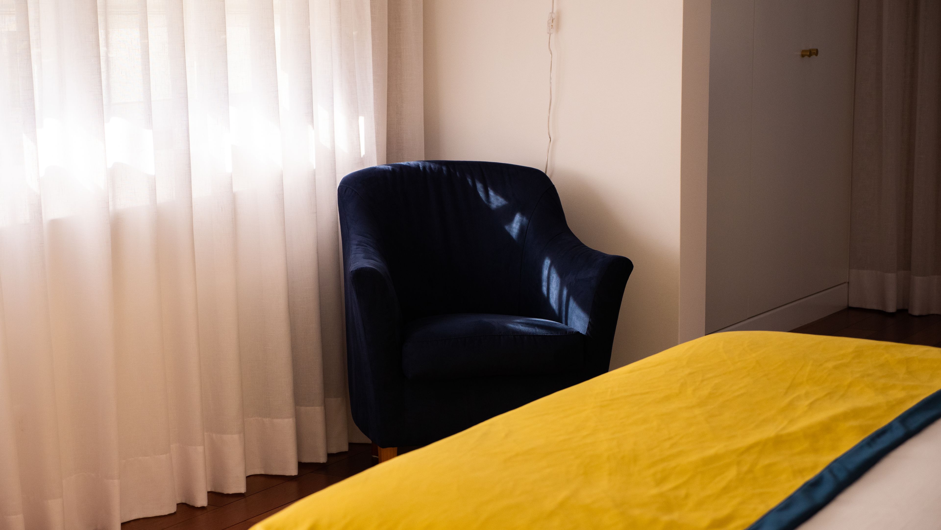 Una habitación con un sillón azul oscuro, una colcha amarilla y cortinas blancas.
