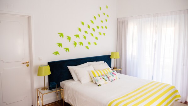 Un dormitorio con cama, dos mesitas de noche, una mesita auxiliar, una ventana con cortinas y una pared decorada con motivos de hojas.