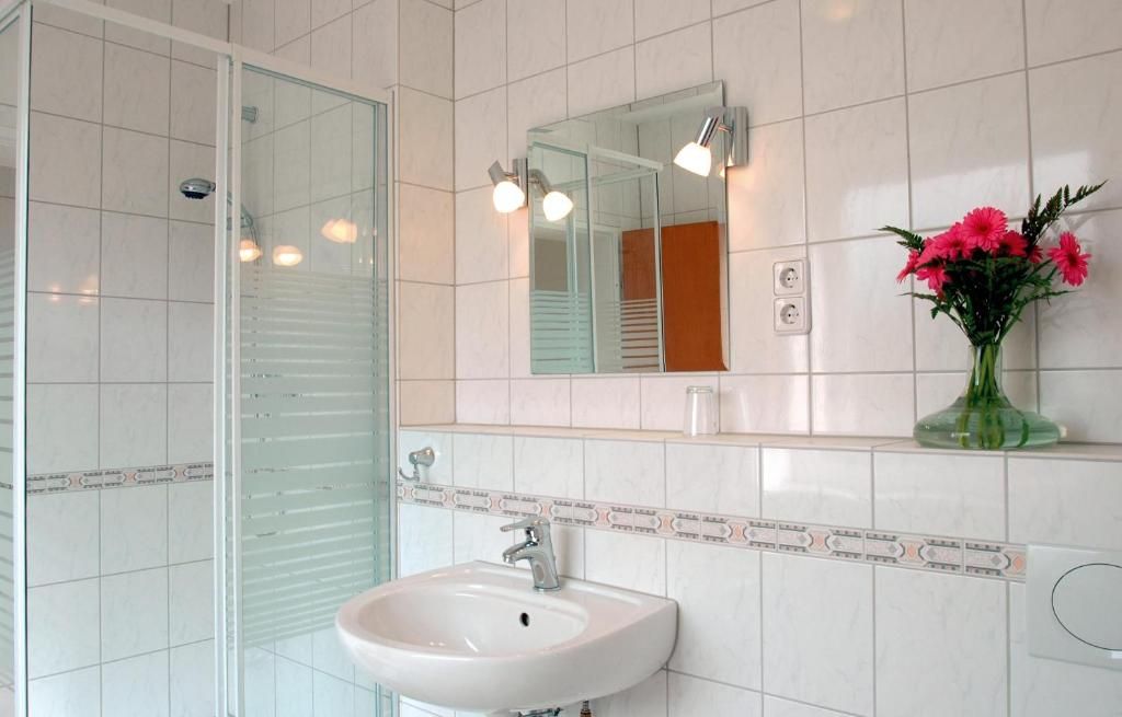 Un baño con cabina de ducha de vidrio, un lavabo blanco, un espejo con luces y un florero con flores en la pared.