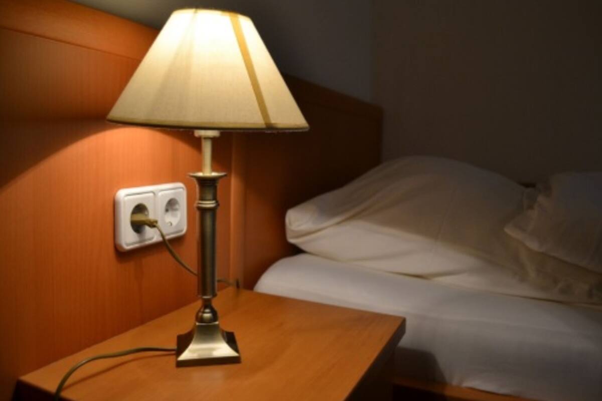 A bedside table with a lamp, a power outlet, and a bed with white bedding.