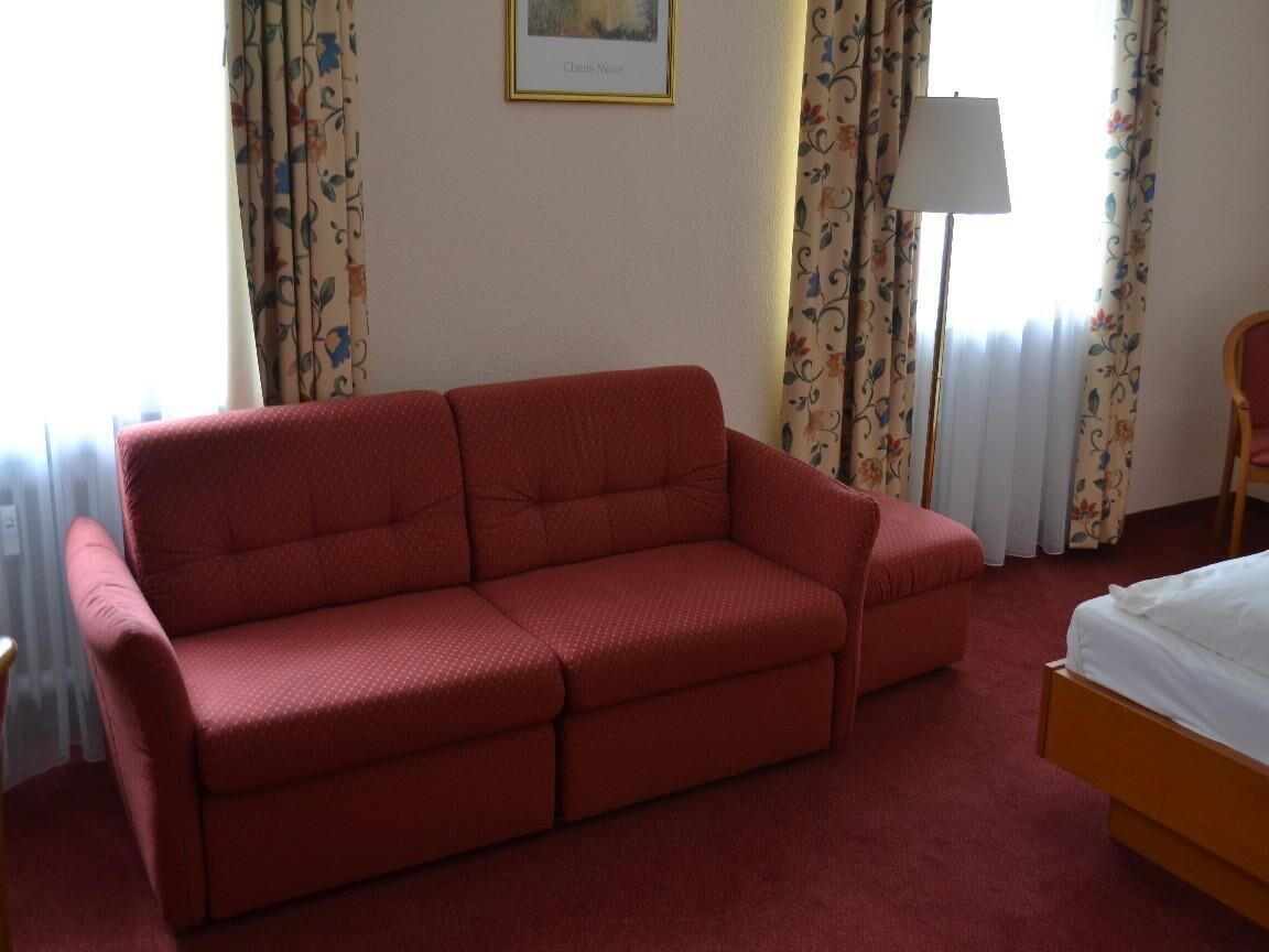 A red sofa in a room with a bed, floral curtains, and a lamp.