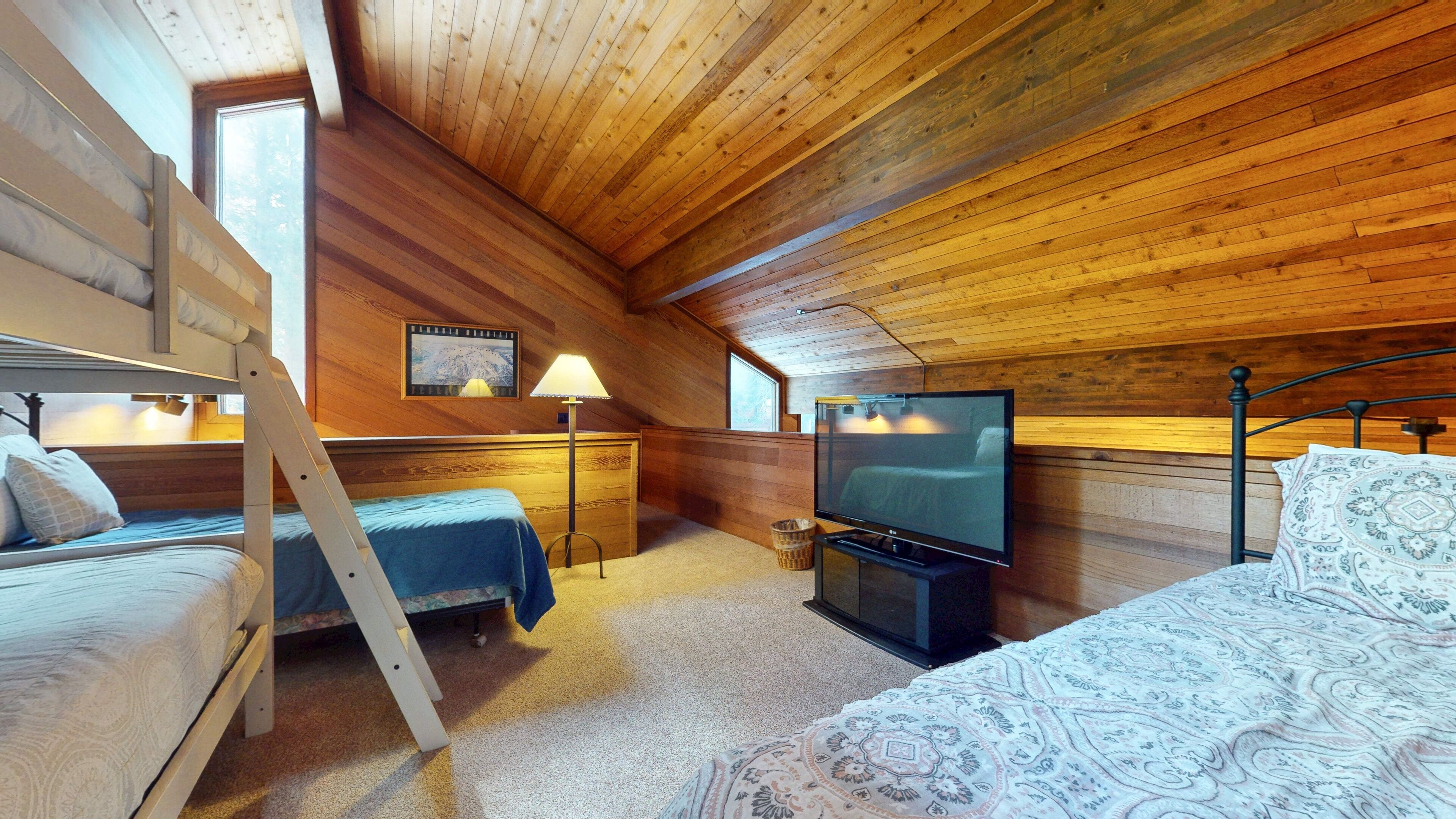 A cozy attic bedroom with wooden walls, a bunk bed, a flat-screen TV, and a desk lamp.