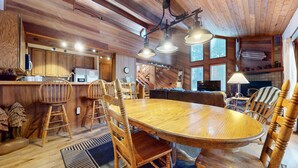 A rustic cabin interior with a wooden dining table, chairs, a kitchen area, and a fireplace.