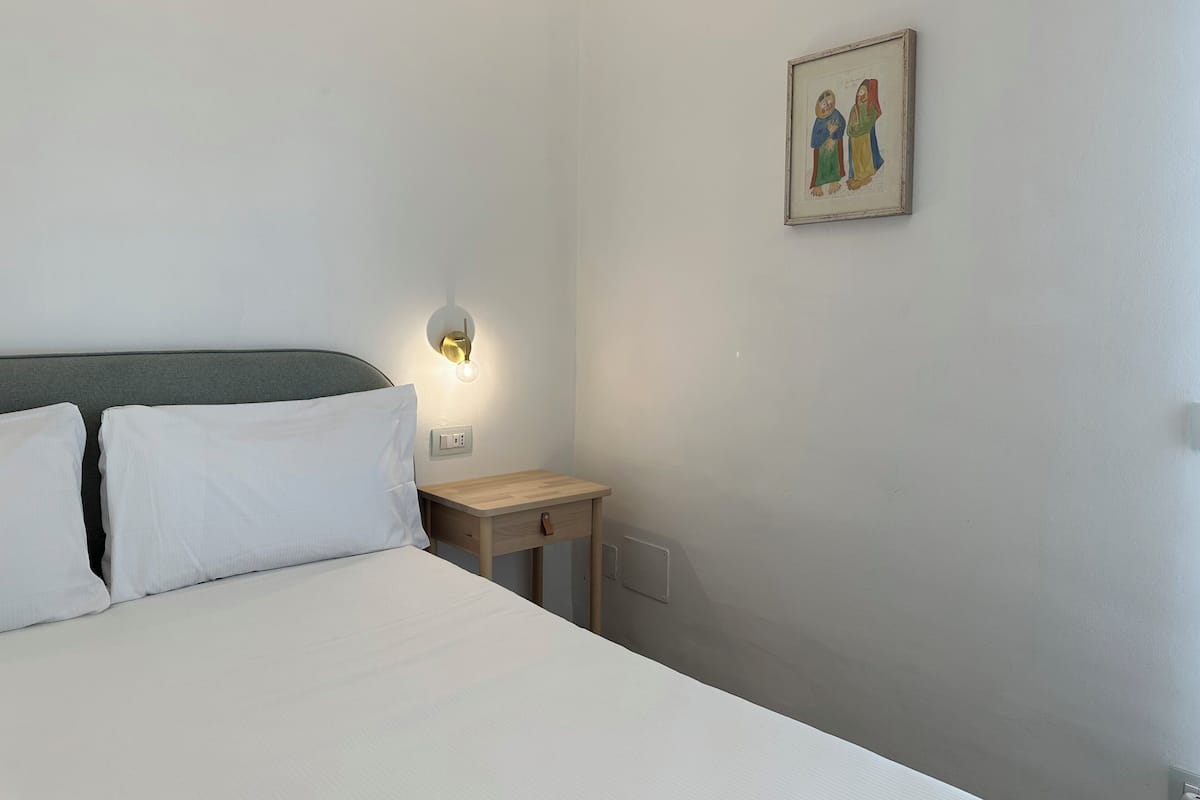 A hotel room with a bed, a bedside table, a wall-mounted light, and a framed picture.