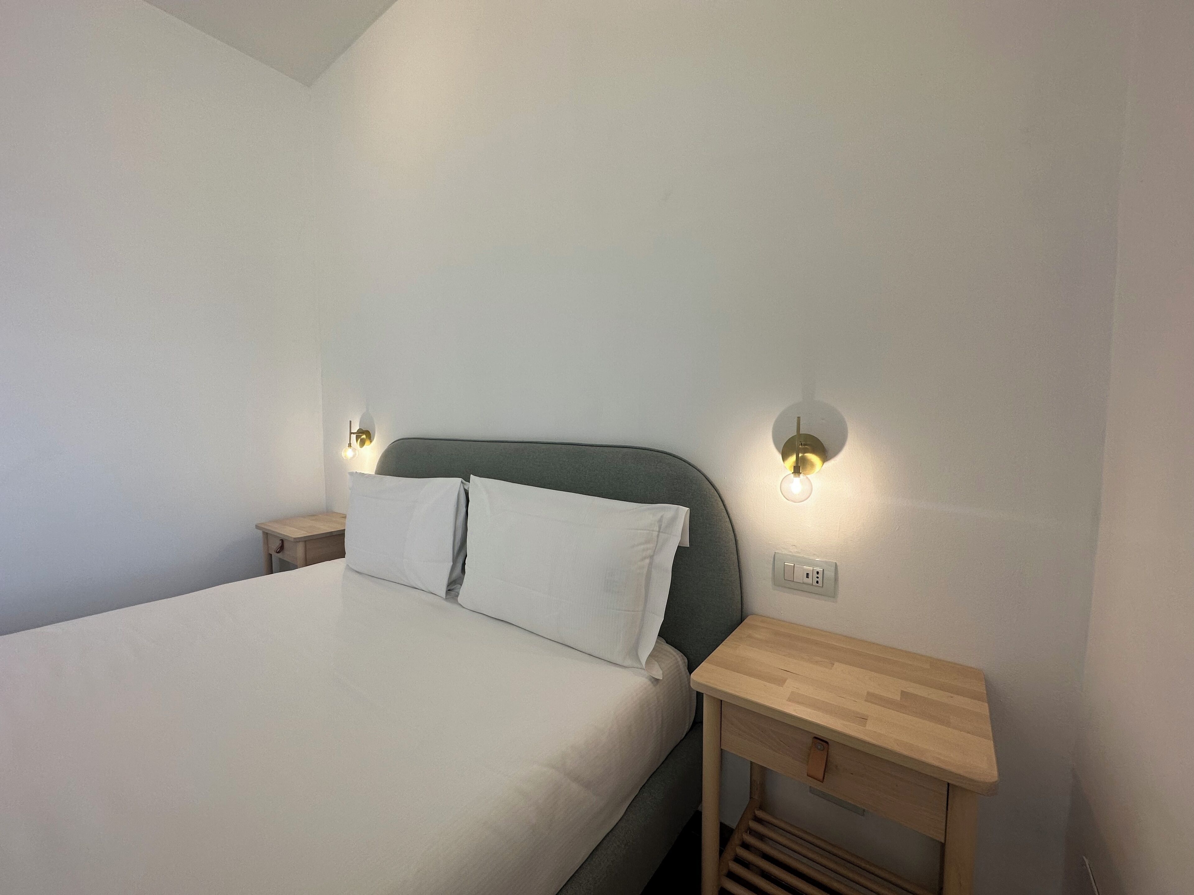 A neatly made bed with white linens, two bedside tables with wall-mounted lamps, and a small wooden nightstand with a drawer.