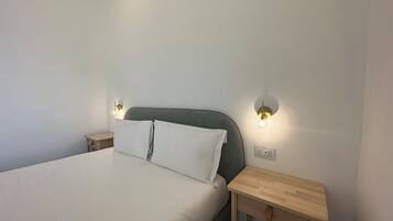 A neatly made bed with white linens, two bedside tables with wall-mounted lamps, and a small wooden nightstand with a drawer.