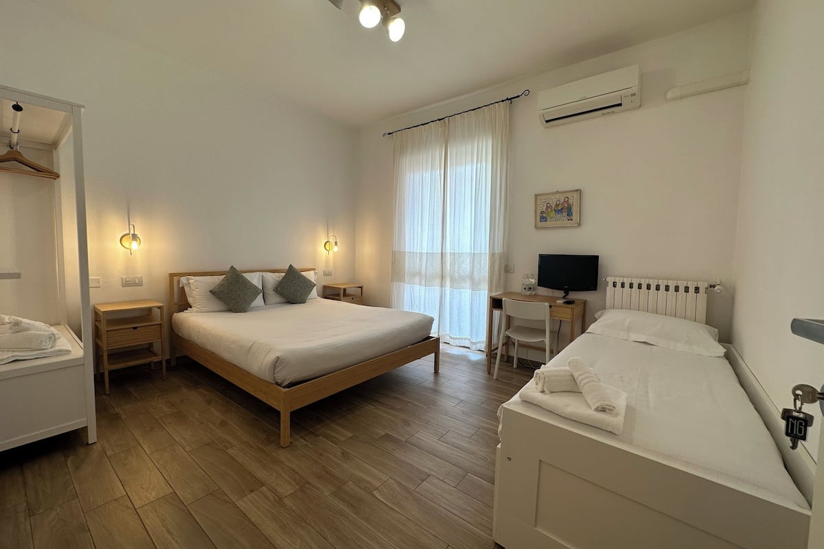 A bedroom with a large bed, bedside tables, a desk with a computer, and an air conditioning unit.