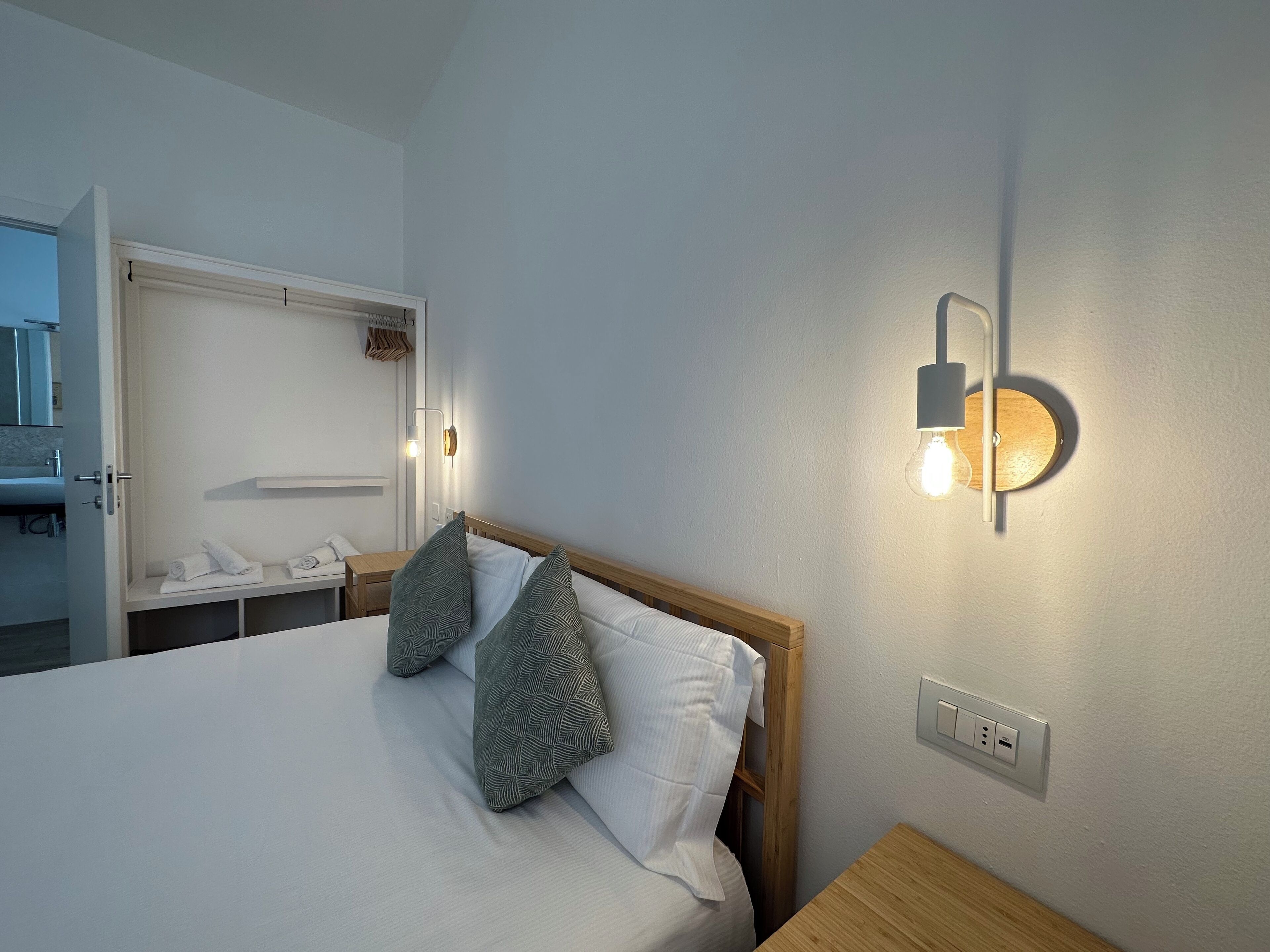 A neatly made bed with two patterned pillows, a bedside table, a wall-mounted light fixture, and a partially visible bathroom with a towel rack.