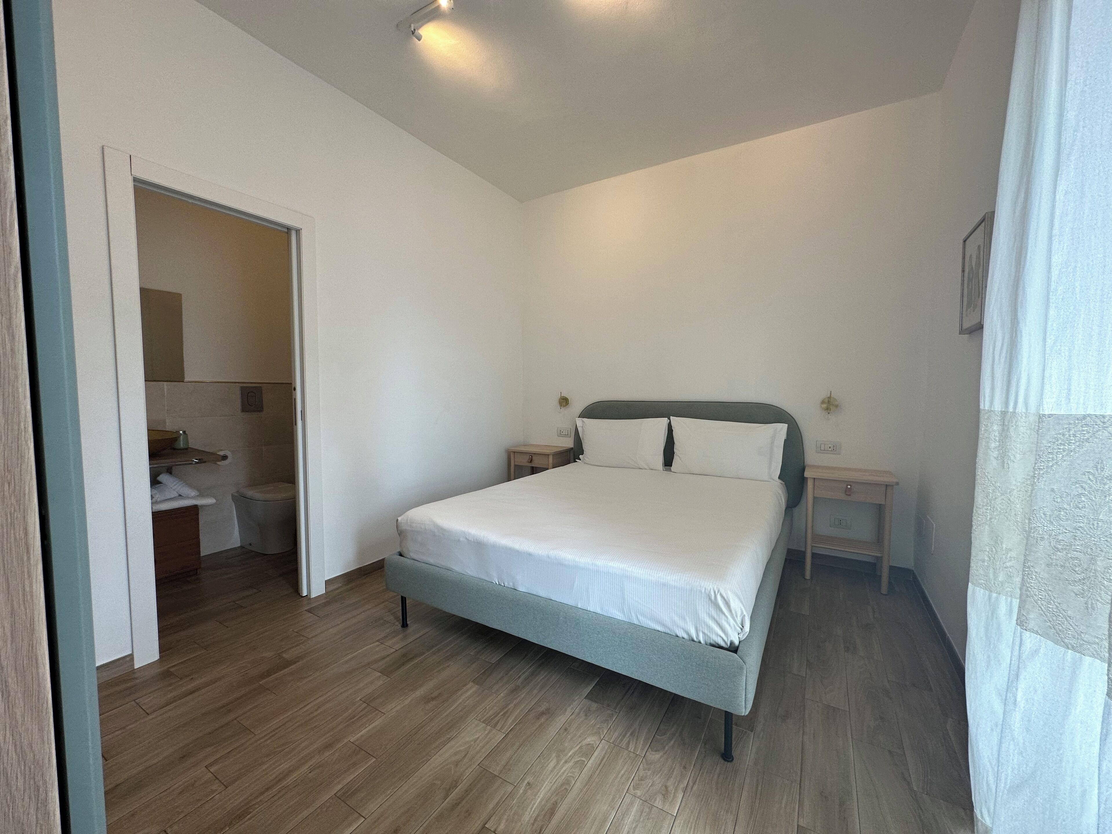 Un dormitorio con cama, mesitas de noche, suelo de madera y una puerta que da al baño.