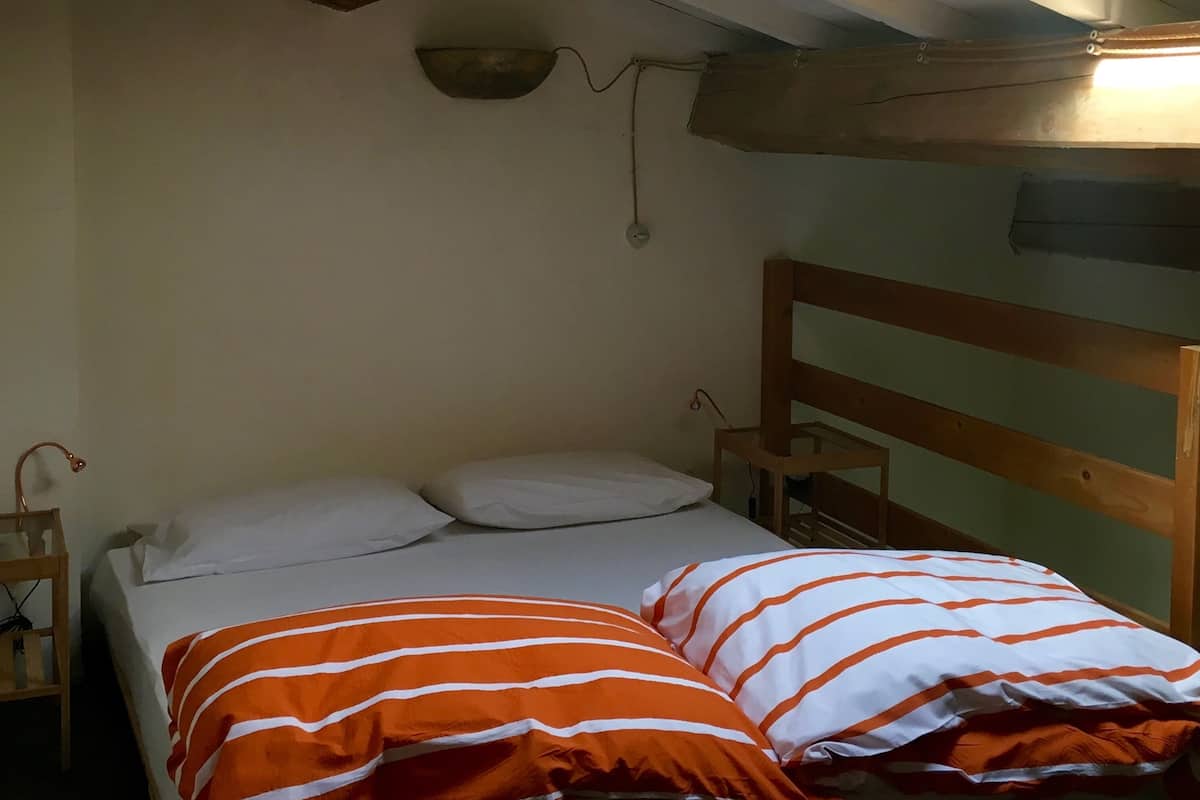 Una habitación pequeña con cama individual, techo inclinado, cabecero de madera y una lámpara fijada a la pared.