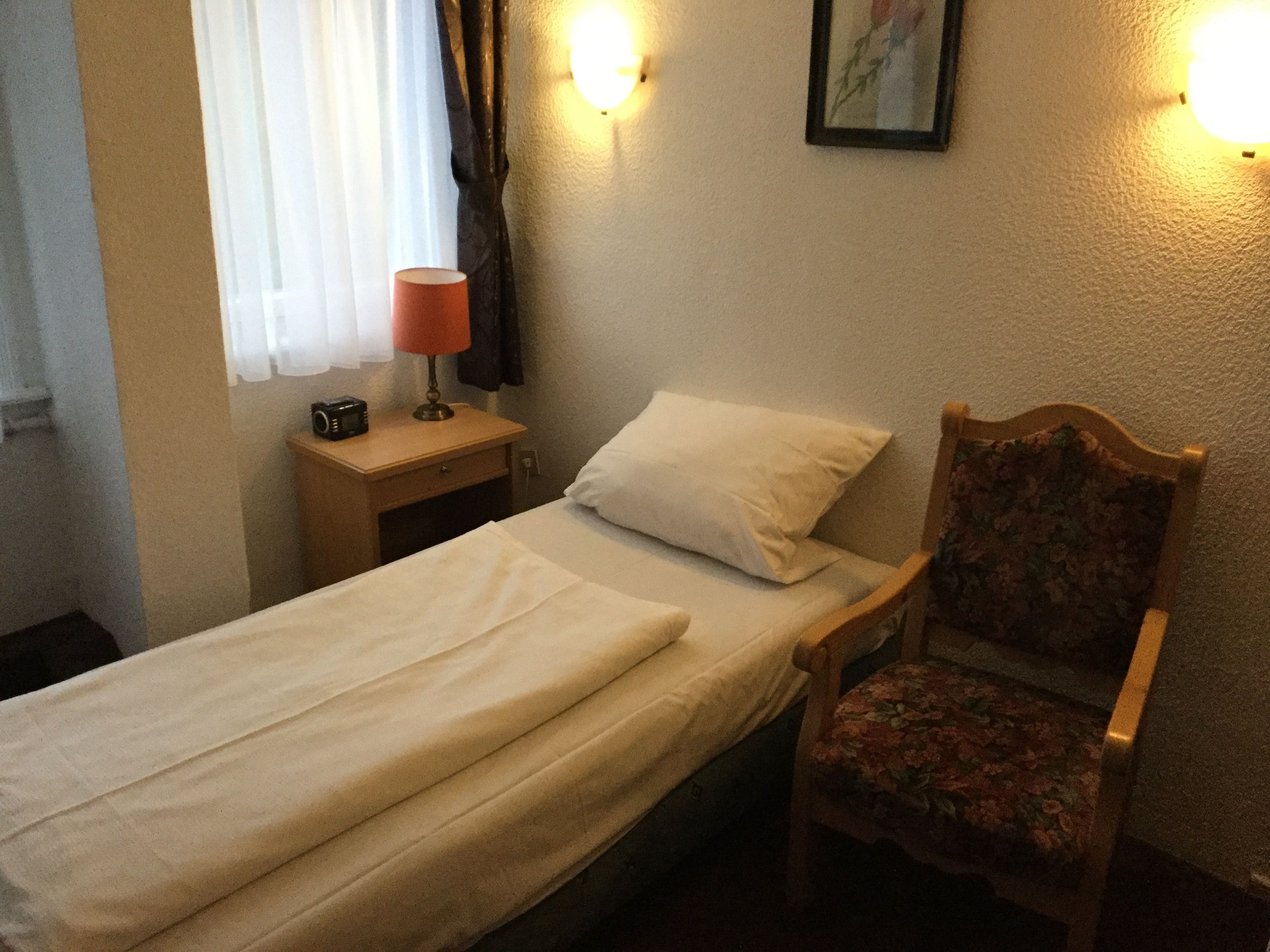 A single bed with a white pillow and a wooden chair with a floral seat. There is a nightstand with a lamp and a camera.