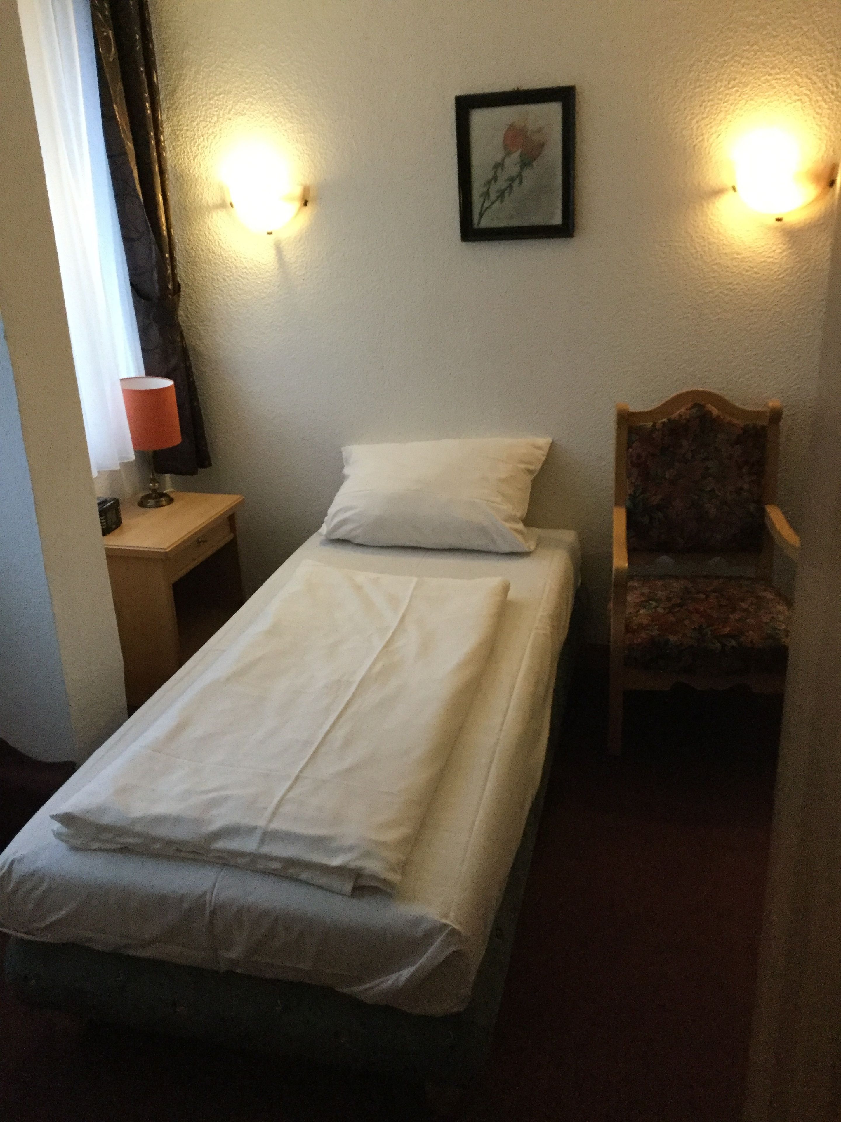 Une petite chambre avec un lit simple, une table de chevet en bois, une chaise et un tableau accroché au mur.