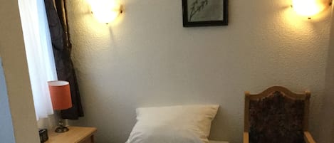 Une petite chambre avec un lit simple, une table de chevet en bois, une chaise et un tableau accroché au mur.