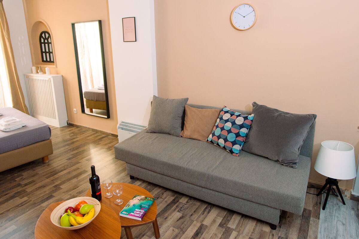 A cozy living room with a grey sofa, a small round table with a bowl of fruit and a magazine, a white lamp, and a clock on the wall.