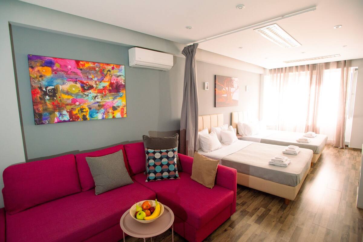 A hotel room with a red sofa, a bed with two pillows, a small table with fruit, and a colorful abstract painting on the wall.