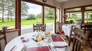 A dining area with a table set for breakfast, large windows offering a view of the outdoors, and a clock on the wall. - Langdon Farm Guest House (Kilgetty)