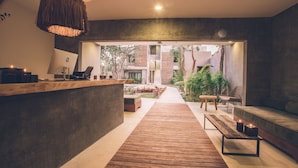 Un interior moderno con un mostrador de recepción de madera, una zona de estar con un banco y un sofá, y vistas a un jardín exterior y un edificio.