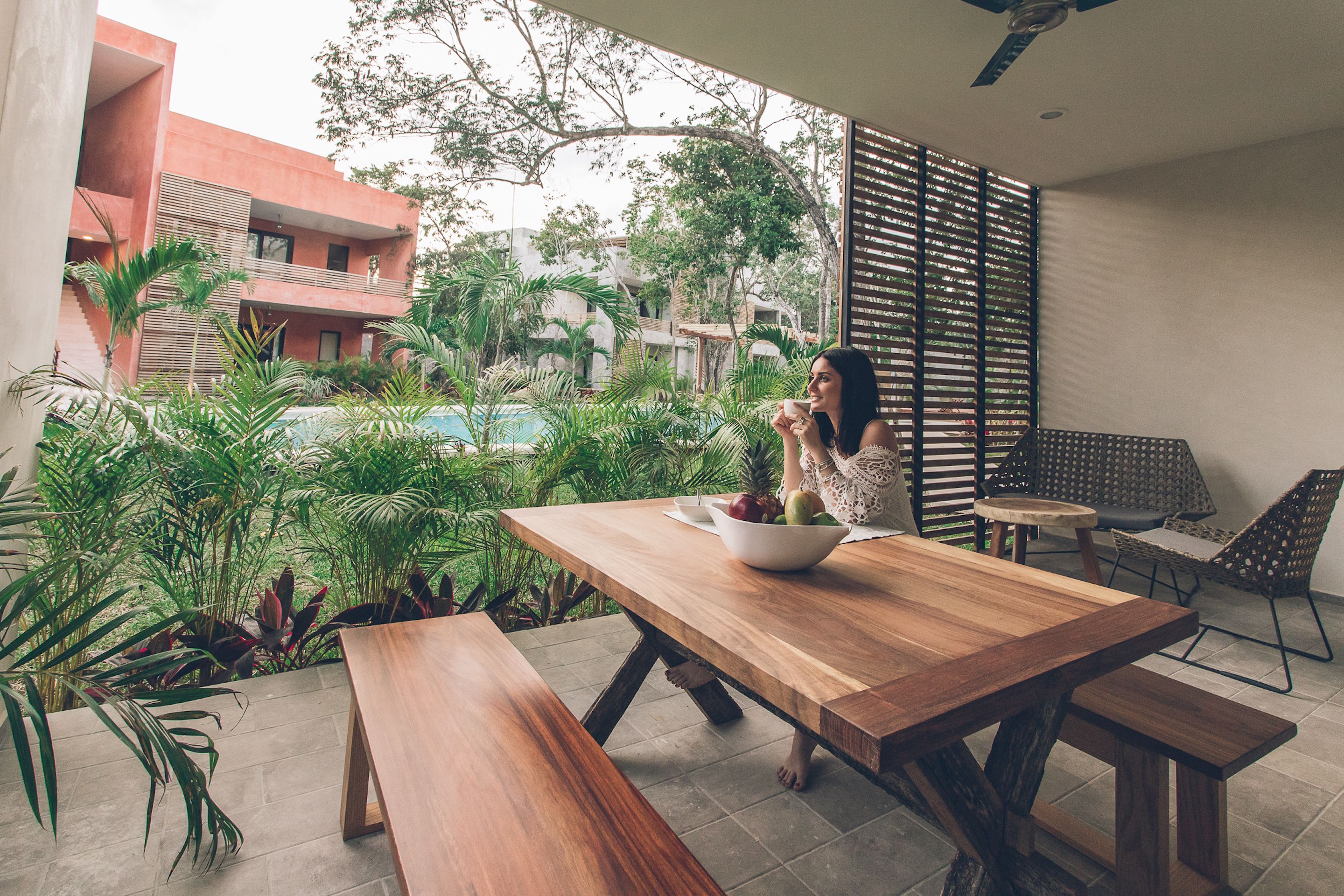 A wooden dining table with a bowl of fruit, surrounded by potted plants and a view of a swimming pool.
