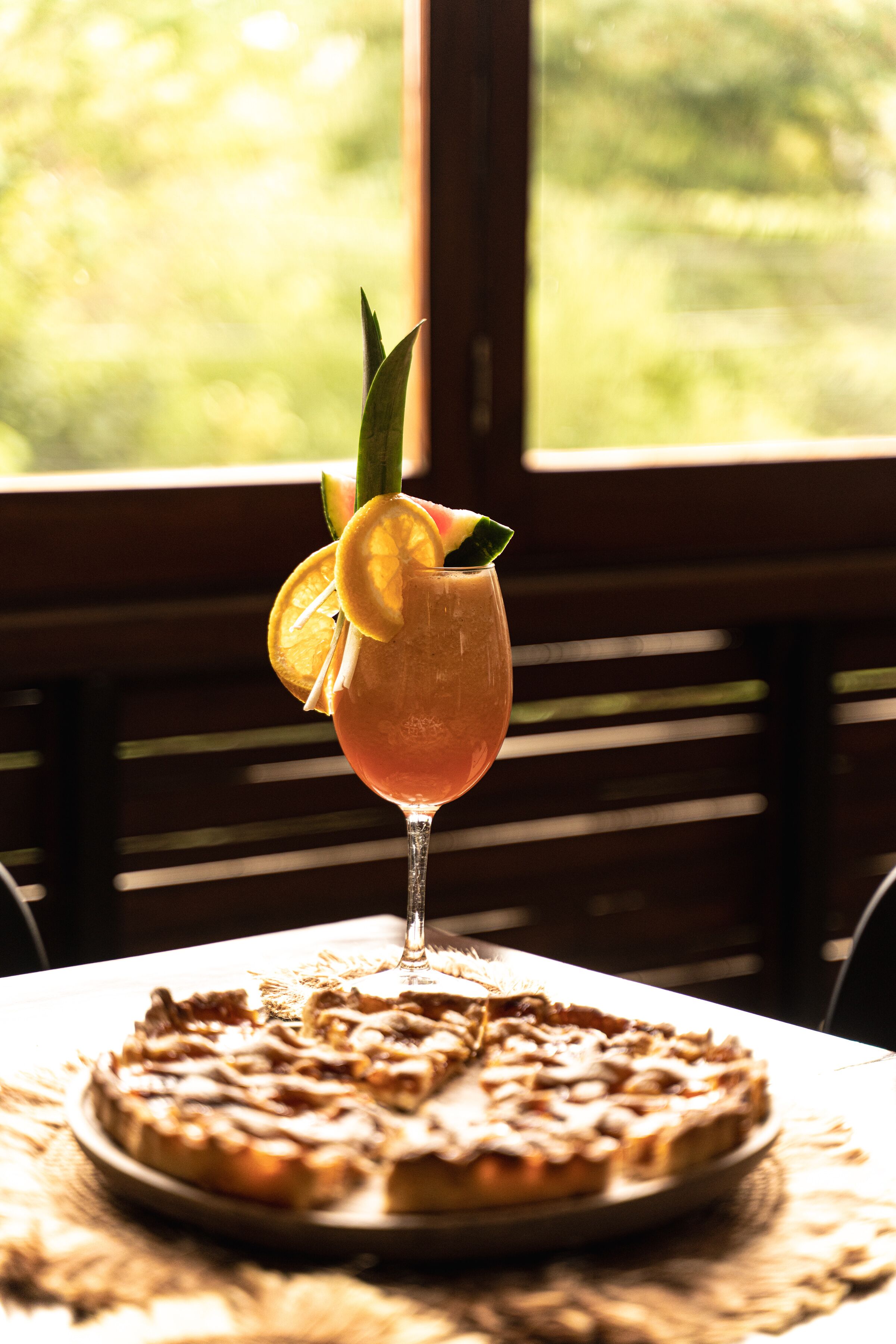 A table with a glass of a beverage garnished with a slice of lemon and a leaf, and a pizza with a white sauce and cheese on a woven placemat.