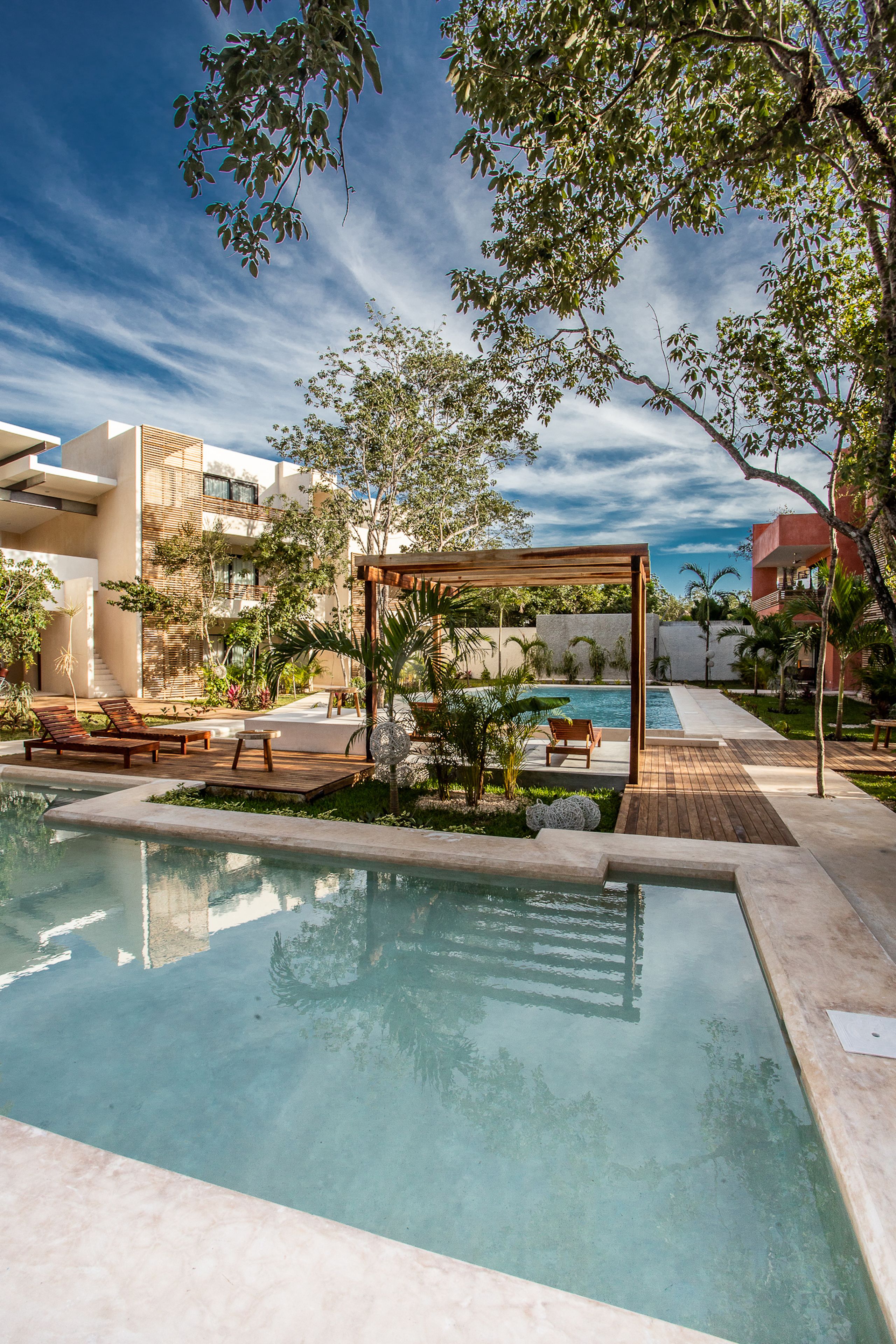 Un espace piscine doté de chaises longues et d’une pergola, entouré de verdure et de bâtiments modernes.