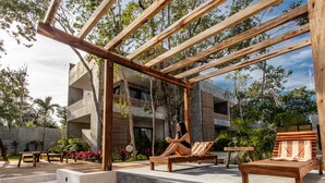 Una terraza de madera con una persona sentada en una tumbona, rodeada de plantas y un edificio moderno al fondo.