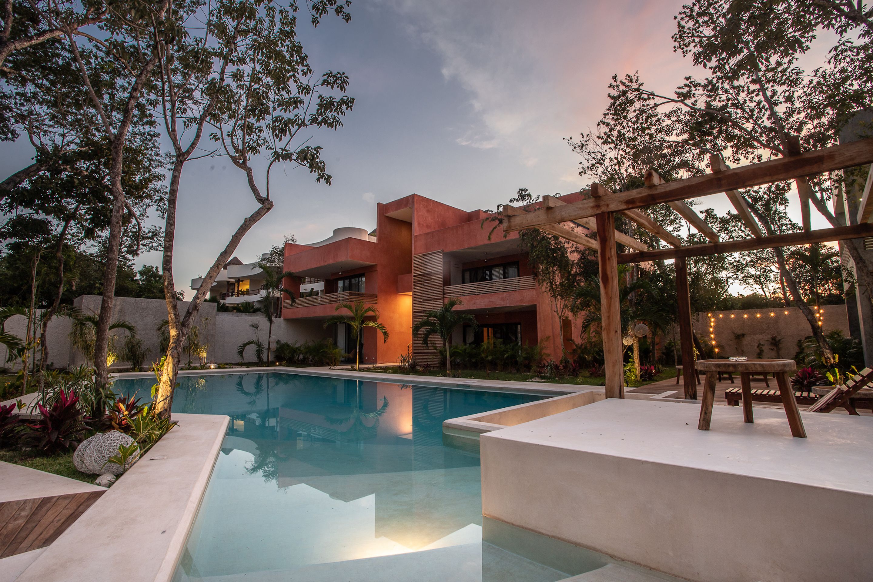 A swimming pool with a wooden deck and lounge chairs, surrounded by lush greenery and a building with a modern design.