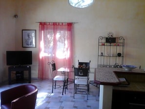Uma sala de estar com televisão, uma mesa de jantar com cadeiras, uma janela com cortinas e uma estante de metal com diversos objetos.