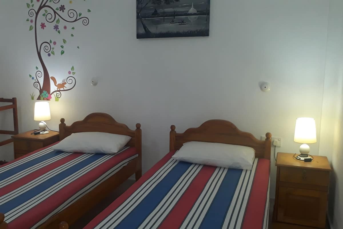 A room with two single beds, each with a striped bedspread and a wooden headboard. There are bedside tables with lamps, a wall decoration of a tree, and a painting above the beds.