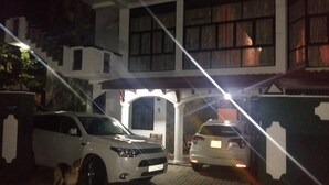 A two-story building with a central entrance, two parked cars, and a dog near the entrance. - Basilico Homestay (Kegalle)