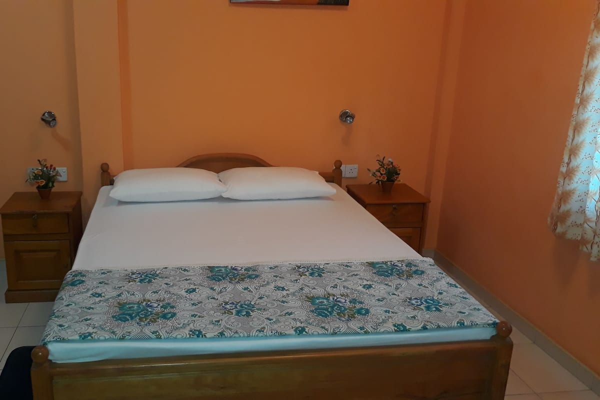 A bedroom with a wooden bed, white pillows, a floral bedspread, two nightstands with small plants, and an orange wall.