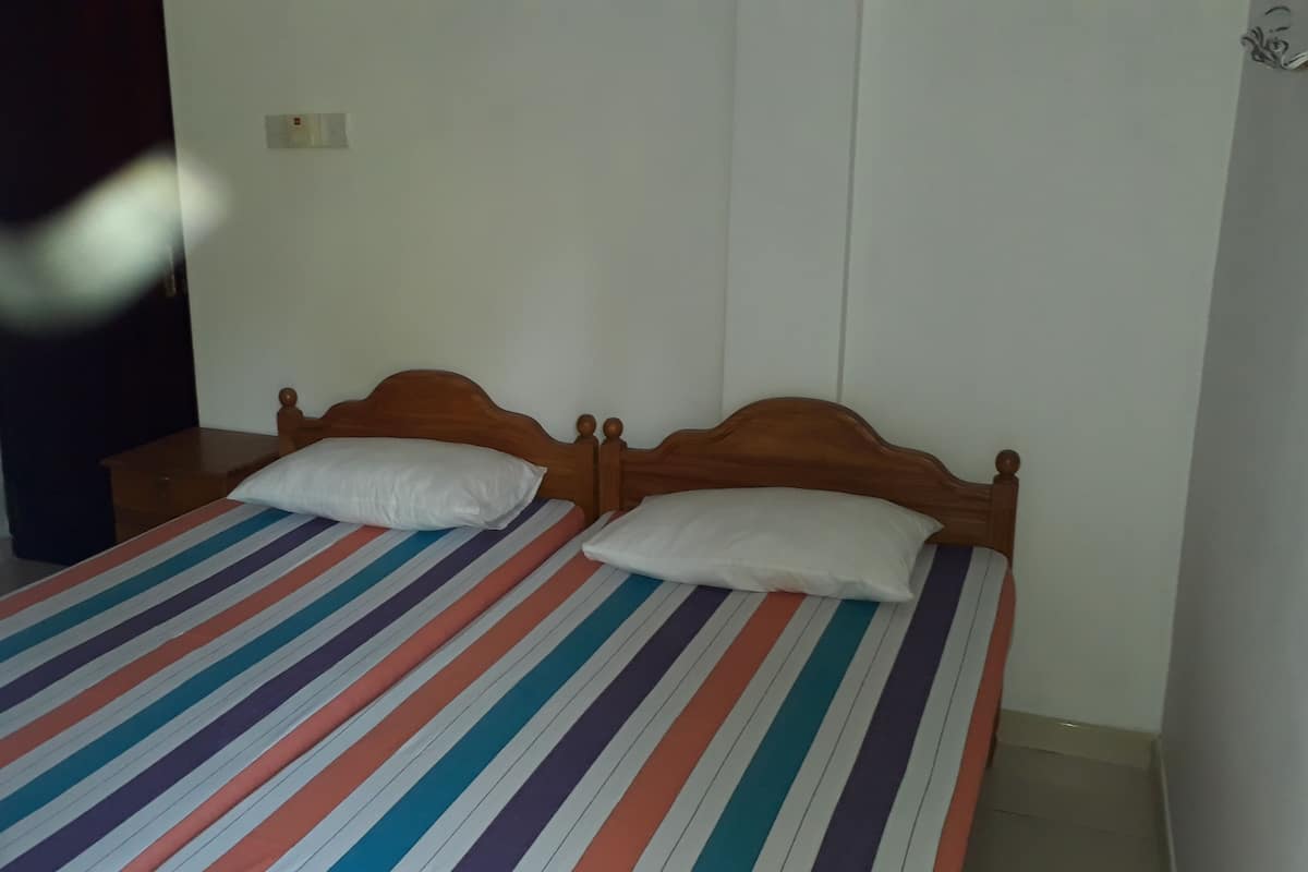 A room with two single beds, each with a striped bedspread and white pillows. There is a wooden bench against the wall and a small table beside the beds.
