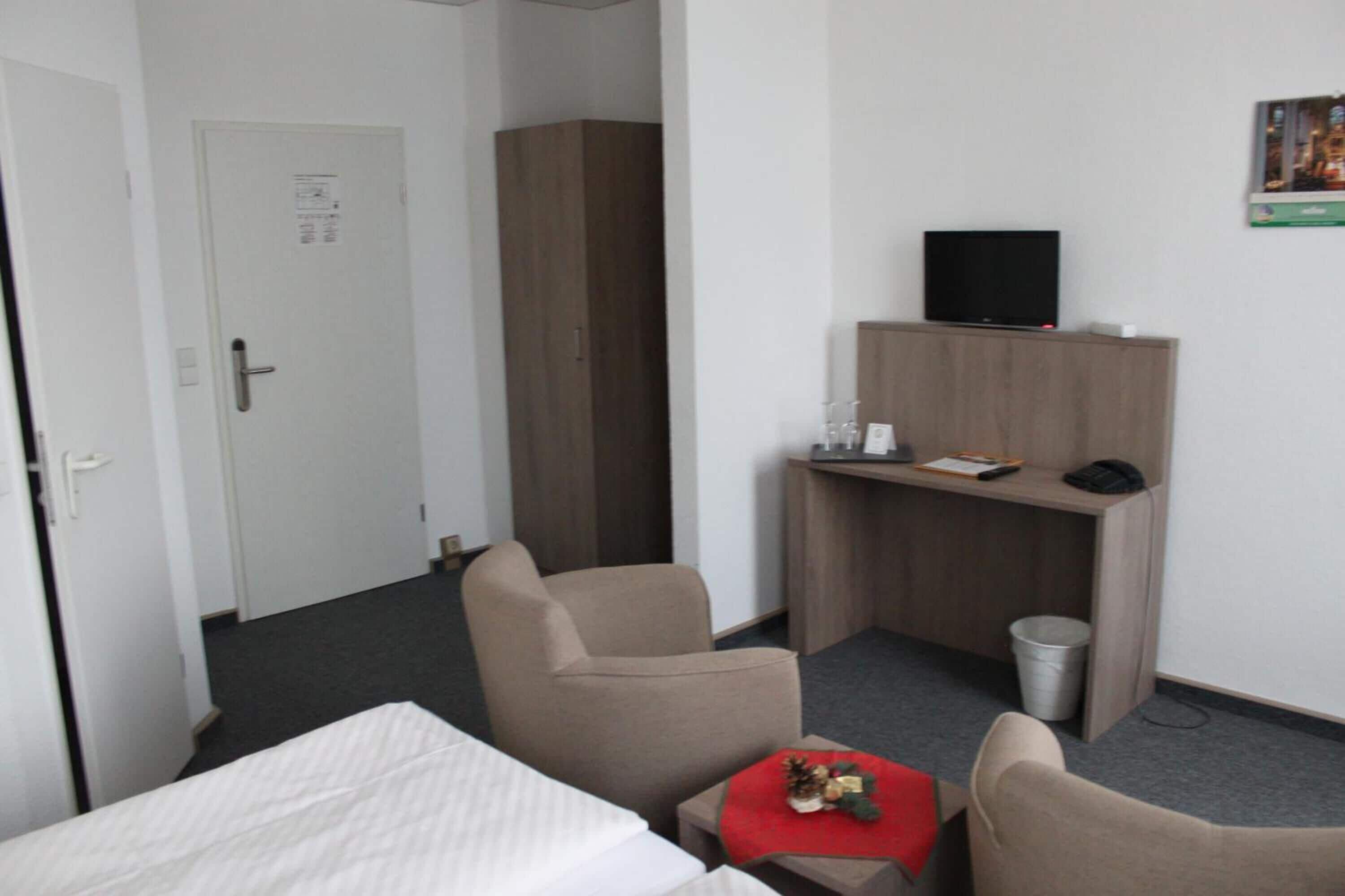 Hotellihuone, jossa on sänky, kaksi tuolia, työpöytä, jossa on televisio, ja roskakori.