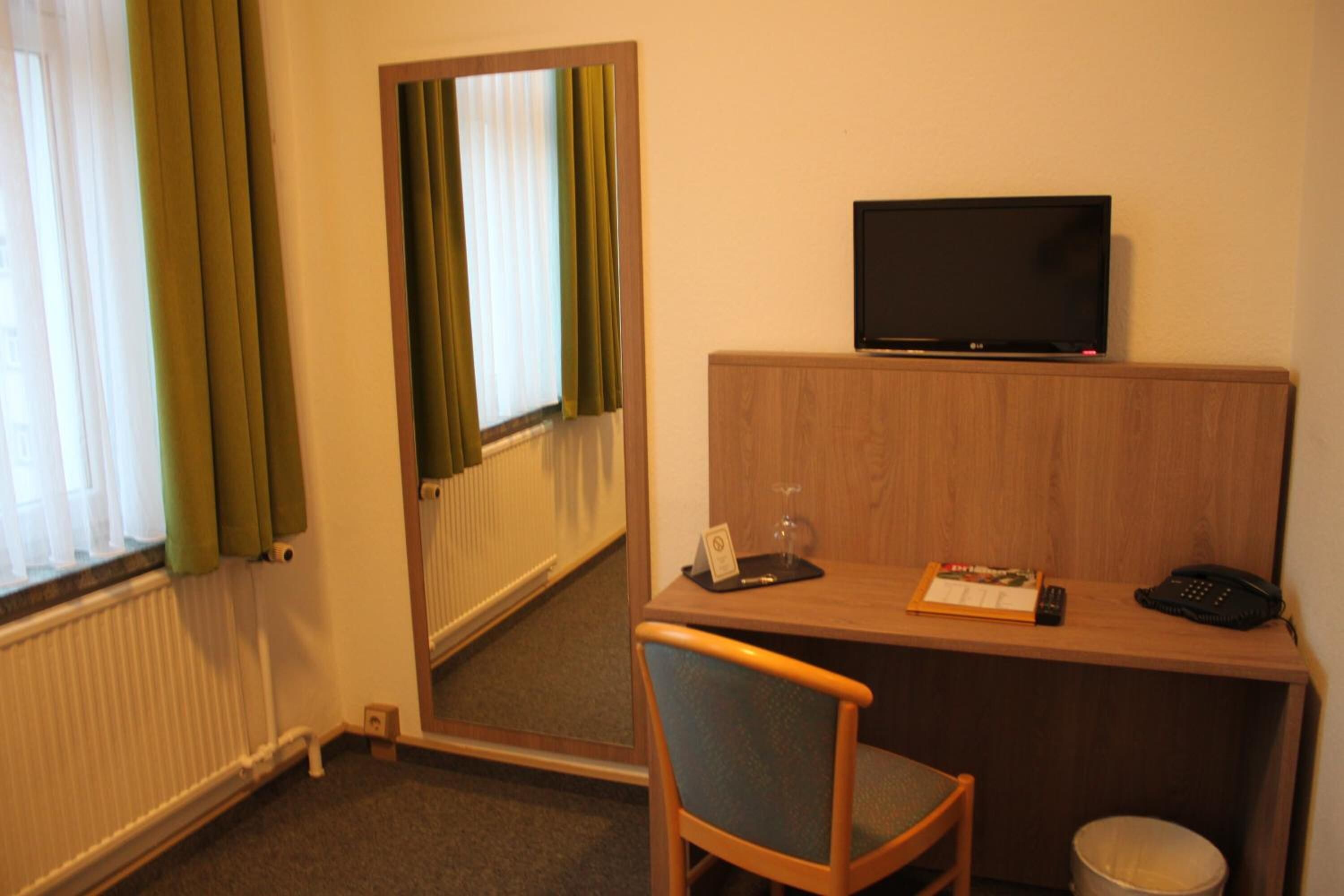 Habitación de hotel con un escritorio de madera, televisión, una silla, un cesto de basura pequeño y una ventana con cortinas.