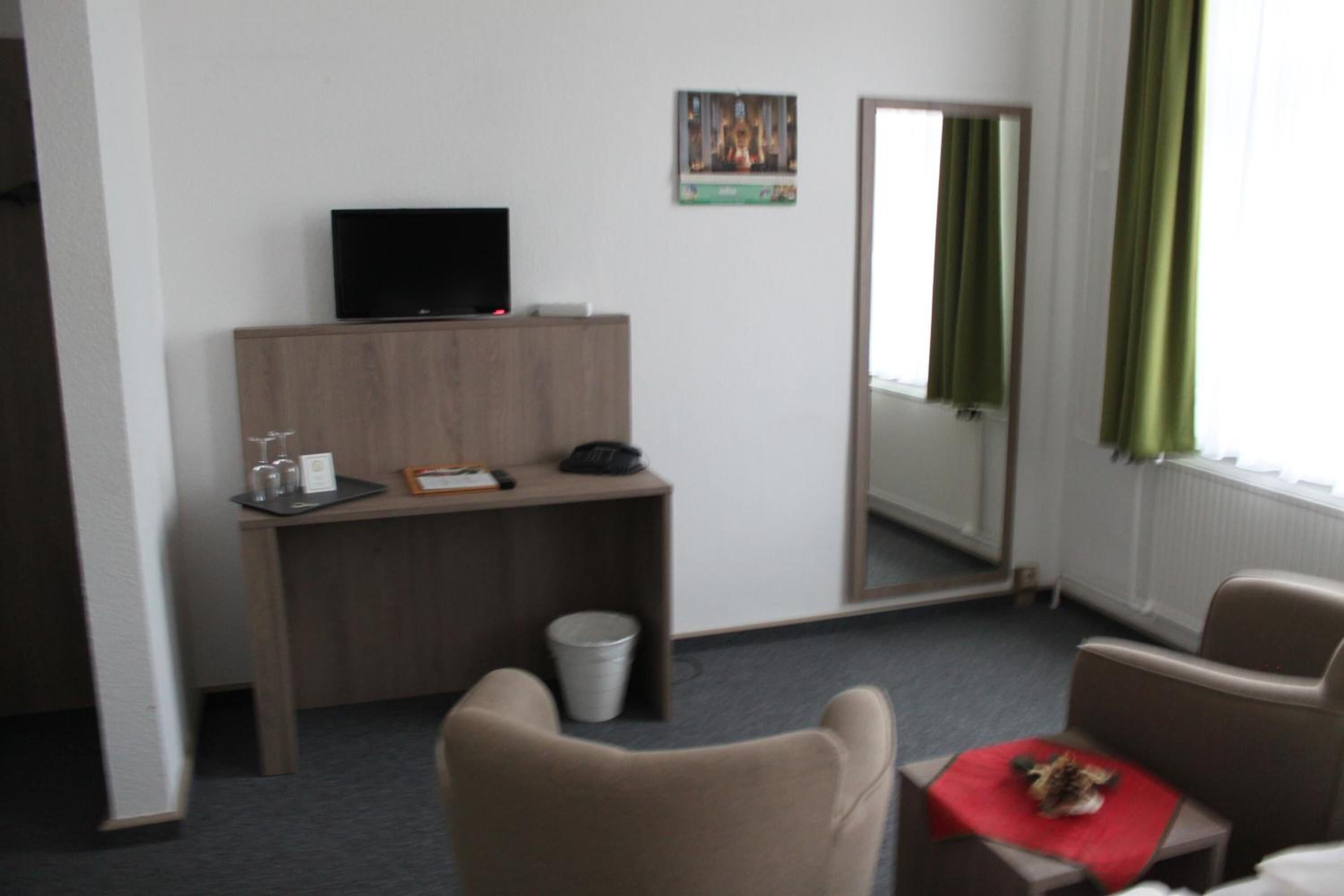 A hotel room with a desk, a television, a chair, a small table with a red cloth, and a mirror reflecting the room.
