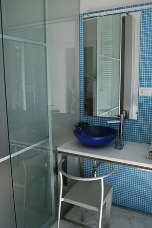 A bathroom with a glass shower enclosure, a blue basin sink, a mirror, and a tiled backsplash. - Hotel Portorais (Carini)